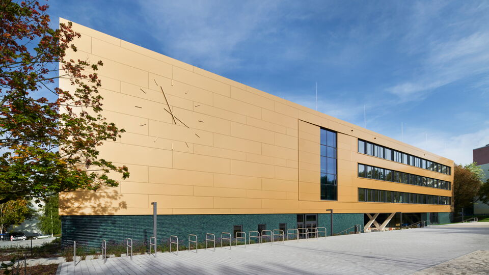 Modernes Gebäude mit goldgelber Fassade und großer Uhr, grünem Sockelbereich, Fahrradständern davor und blauem Himmel.